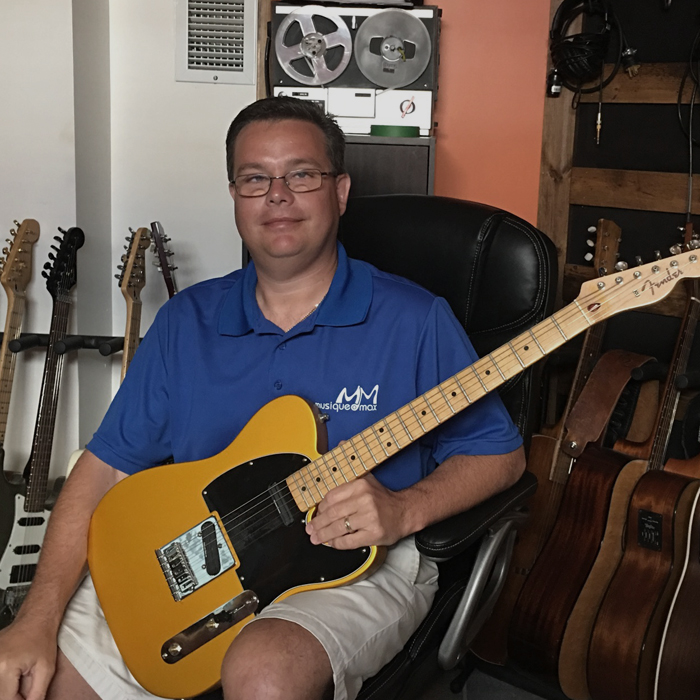 Tommy Théberge assis avec une guitare à la main et portant un chandail bleu avec le logo de Musique O Max.