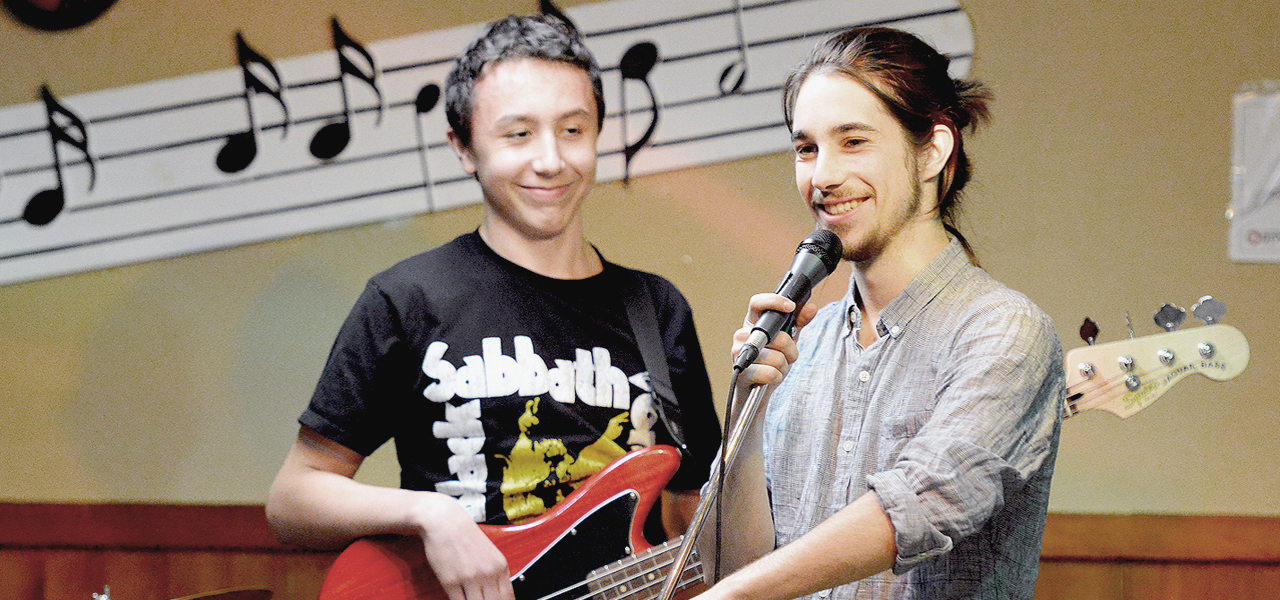 Un adolescent souriant jouant de la basse accompagné de son professeur.